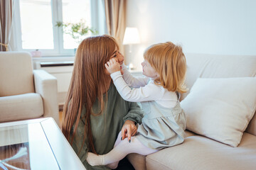 Obraz premium Mother and daughter having fun on sofa. Beautiful young woman and her charming little daughter are hugging and smiling. Happy loving family. Mother and her daughter child girl playing and hugging.