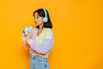 Asian girl in headphones holding instant camera isolated over yellow background