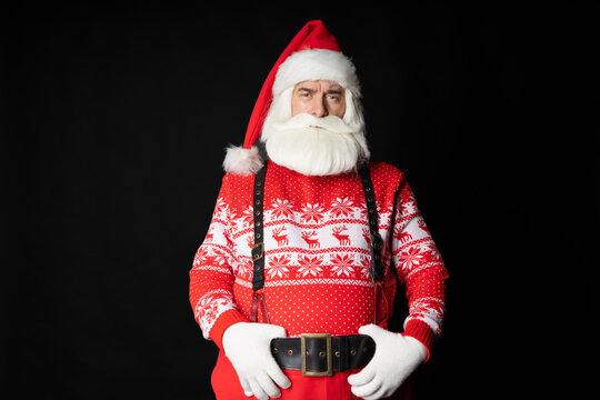 Photo of an elderly Santa in a sweater, holding his hands on a black belt with suspenders, a businesslike look of a worker in a helmet. Portrait on a black background.