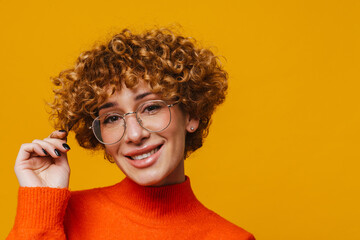 Middle-aged woman in glasses touching hair isolated over yellow background