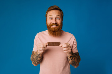 White bearded man eating chocolate bar isolated on blue background