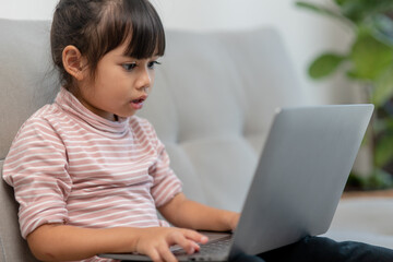 Asian Little school kid girl use laptop computer sitting on sofa alone at home. Child learning reading online social media content, play education lessons game chatting with friends. 