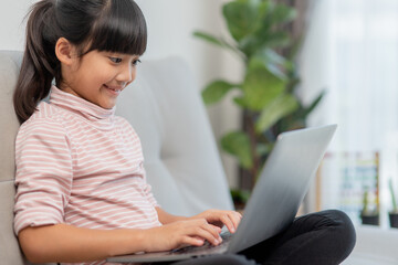 Asian Little school kid girl use laptop computer sitting on sofa alone at home. Child learning reading online social media content, play education lessons game chatting with friends.