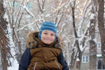 Portrait of an attractive boy outdoors in winter. He is wearing a warm hat and jacket, looking at the camera, and smiling. Against the background of trees in the snow and a multi-story building.