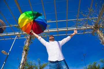 Handsome young blue eyed gay man with open arms holding a rainbow coloured umbrella in his hand. The photo is taken from below and you can see the blue sky in the background.
