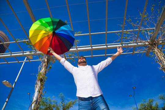 Handsome young blue eyed gay man with open arms holding a rainbow coloured umbrella in his hand. The photo is taken from below and you can see the blue sky in the background.