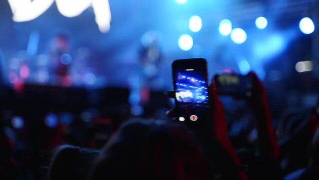 Close up of man hands silhouette taking photo or recording video of live music concert with smartphone at open air festival. Bright colorful stage lighting. Fans at favourite band performance.