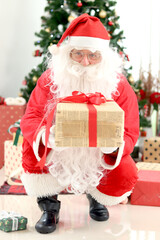 Happy smiling Santa Claus holding gift box under Christmas tree in white living room , Santa giving present to camera, celebrating happy Christmas winter holiday