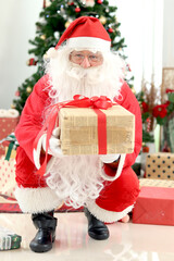 Happy smiling Santa Claus holding gift box under Christmas tree in white living room , Santa giving present to camera, celebrating happy Christmas winter holiday