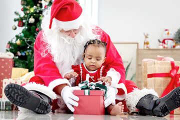 Fototapeta na wymiar Adorable happy smiling African American child girl sitting on Santa Claus lap around decorative Christmas tree, kid open Christmas gift box present, feeling surprised and excited