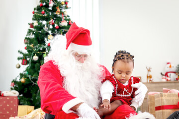 Fototapeta na wymiar Adorable happy smiling African American child girl sitting on Santa Claus lap around decorative Christmas tree, kid open Christmas gift box present, feeling surprised and excited