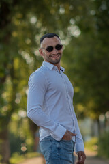 Attractive young man, wearing a white shirt and sunglasses, looks at the camera and smiles. a stylish guy in a white shirt and jeans walks in the city, a businessman at lunch