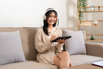 Female entrepreneur is sitting on big comfortable sofa and wearing headphone while holding tablet to watching