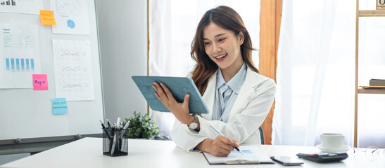 Businesswoman in suit reading business data on tablet and taking notes information in paperwork while working