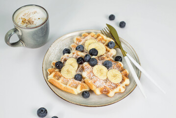Homemade viennese waffles with blueberries and banana on a light plate on a light background.