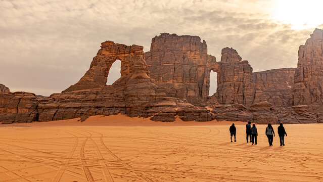 Tamesguida cathedral in Tadrart Rouge. Walking tourists. Five silhouettes with shadows walking away, orange color sand with sunny cloudy sky. Naturally sculpted rocky mountains of the Tassili N'Ajjer