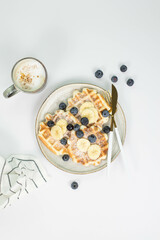 Homemade viennese waffles with blueberries and banana and cup of coffee on a light background.
