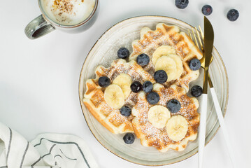Homemade viennese waffles with blueberries and banana and cup of coffee on a light background.