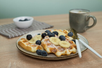 Homemade viennese waffles with blueberries and banana and cup of coffee on wooden.