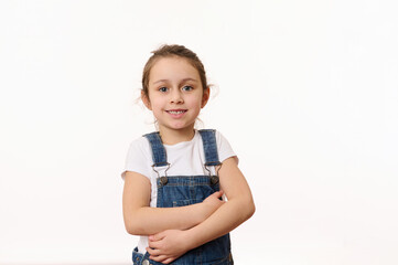Emotional portrait of a Caucasian cute baby girl, wearing blue denim overalls, happily smiling looking at camera, hugging herself while posing over white background with copy ad space for promo text