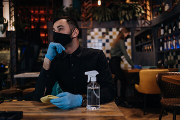 Young restaurant waiters cleaning and disinfecting tables and surfaces during Coronavirus pandemic disease. They are wearing protective face masks and gloves.