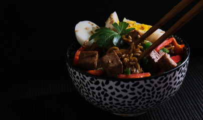 Cuenco de noodles asi&aacute;ticos con tofu, huevo y verduras en fondo negro. Darck food
