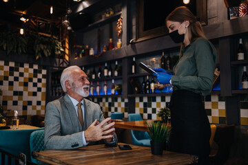 Waitress serves and takes the order from the senior businessman at the restaurant. She wears a protective mask as part of security measures against the Coronavirus pandemic.
