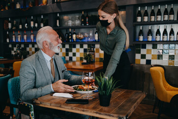 Waitress serves and takes the order from the senior businessman at the restaurant. She wears a protective mask as part of security measures against the Coronavirus pandemic.
