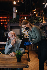 Waitress serves and takes the order from the senior businessman at the restaurant. She wears a protective mask as part of security measures against the Coronavirus pandemic.