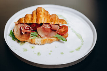 Croissant sandwich with prosciutto, tomatoes, cheese parmesan and greens on black shale board over black stone background. Healthy food concept, top view