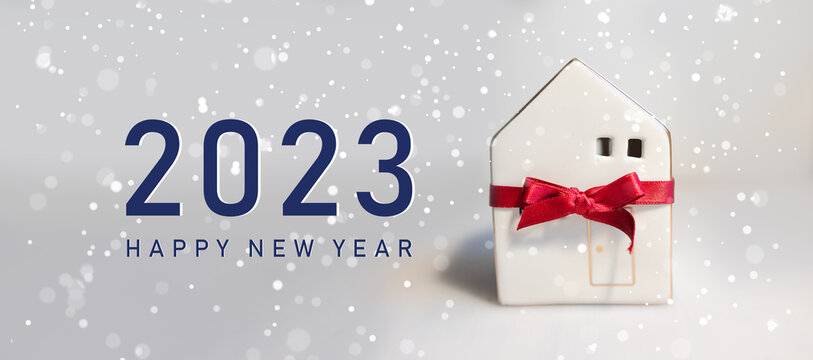 Merry Christmas and 2023 happy new year horizontal banner with small toy model house wrapped in red satin ribbon on a white background. Miniature white toy house model on color background.