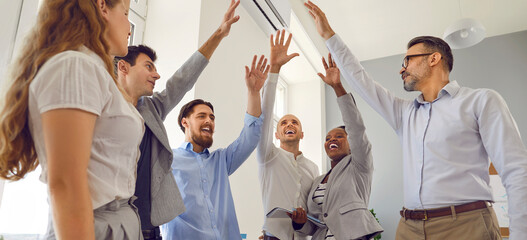 Fototapeta na wymiar Happy cheerful business team celebrating success and having fun at work. Multiracial group of joyful smiling confident people standing in office and raising hands up together. Teamwork concept banner