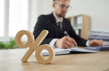 Close up of wooden percent sign on table as symbol of corporate tax and interest rate. Percent sign standing on background of serious busy man in suit calculating on calculator. Blurred background. - obrazy, fototapety, plakaty