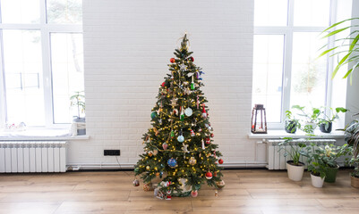 Christmas tree in the white interior of a house with large windows. Glowing fairy lights garlands interior decoration of the studio room. Potted plants in the home