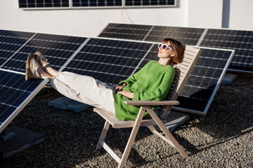 Young woman sits relaxed on a lounge chair while resting on rooftop with a solar station. Happy owner of energy-independent household. Eco-friendly and sustainable lifestyle - 548843349