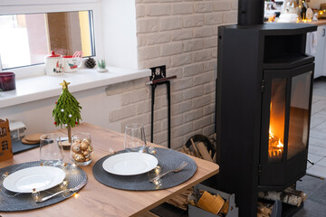 Festive interior of house is decorated for Christmas and New Year in loft style with black stove, fireplace, Christmas tree. Warm studio room with white kitchen, burning wood, cozy and heating of home