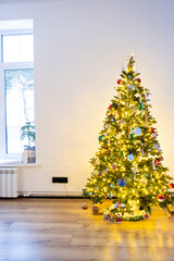 Christmas tree in the white interior of a house with large windows. Glowing fairy lights garlands interior decoration of the studio room. Potted plants in the home