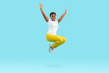 Fototapeta na wymiar Full body length happy cheerful positive crazy excited young African American woman in white T shirt, yellow pants and sunglasses enjoying weekend or holiday mood and jumping in air on blue background
