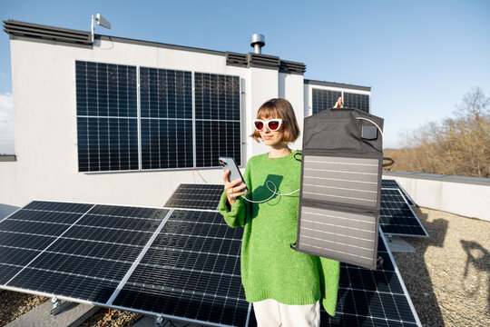 Young woman charging phone from a solar portable panel while standing on rooftop with a solar power plant on it. Concept of alternative energy and new technologies