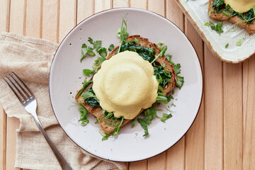 Vegan eggs benedict dish with sourdough bread, creamy plant-based hollandaise sauce, smoked tofu, young onion, parsley, greens, pea sprouts.