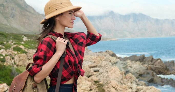 woman tourist in safari hat stands near the ocean. Hipster young girl with backpack enjoying sunset on peak of foggy mountain. Traveler walking along the road to the mountains. Hiking in mountains.