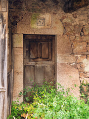 Puerta de madera marrón, de una casa abandonada con hierbas verdes delante, y fachada de piedras, en un pueblo de Asturias, España, en verano de 2021.