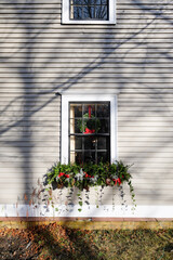 Fototapeta na wymiar A garland of green ivy, fir tree branches, red ribbon, silver Christmas decorations, and red berries hanging under a vintage casement glass window. The exterior wall is tan in color with white trim. 
