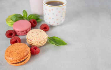 Three multi-colored macaroons, macarons, raspberries and two cups on a gray background with copy space. French dessert and red berries. Lunch