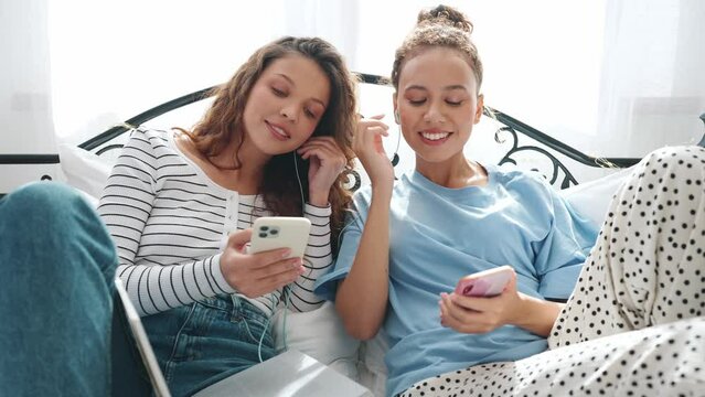 Lovely girlfriends typing on phones and listening music in headphones on the bed 