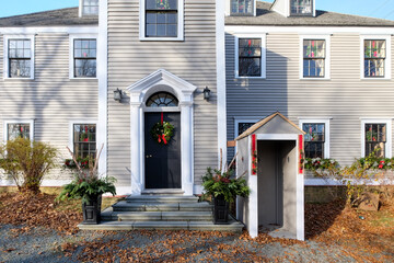 A black exterior single wooden door with a Christmas wreath hanging on the entrance. There are two holiday arrangements on the steps to the vintage tan colored house with red ribbons and garland.