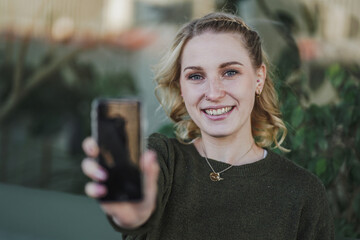 happy smiling young natural alternative looking blond woman in an optimistic & cheerful mood holding or showing mobile phone into the camera pointing to the front with green plants in the background