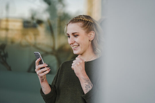 happy smiling young hipp looking blond girl full of joy in a cheerful mood holding and looking into her mobile phone with green plants and glass wall in the background