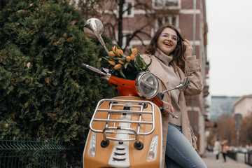 Fototapeta na wymiar Young beautiful hipster girl riding on motorbike city street, autumn europe vacation, traveling, romance, smiling, happy, having fun. Beautiful young girl with scooter. Girl scooter with flower.
