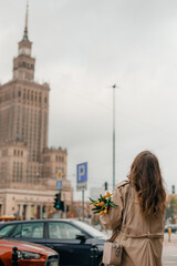 Fototapeta na wymiar A young girl in glasses with flowers against the background of the Palace of Culture in Warsaw. A pretty girl in glasses with flowers walks in Warsaw.